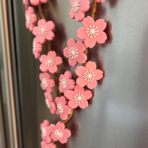 May include: A decorative floral branch with pink cherry blossom flowers and white centers. The branch is brown and appears to be made of wood. The flowers are attached to a metallic surface, possibly a refrigerator.