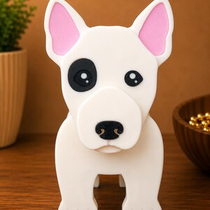 May include: A white dog figurine with pink inner ears and a black eye patch. The dog has a black nose and is standing on four legs. The figurine is made of a smooth material and is set against a brown background.
