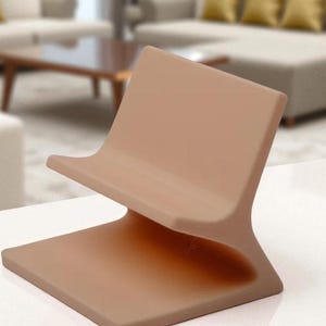 May include: A contemporary phone stand in a warm beige hue. The stand has a curved back and a flat base, designed for a phone or tablet. The stand is made of a smooth, solid material. The design is minimalist.
