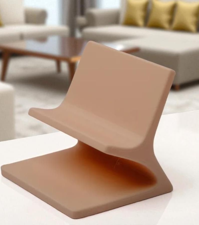 May include: A contemporary phone stand in a warm beige hue. The stand has a curved back and a flat base, designed for a phone or tablet. The stand is made of a smooth, solid material. The design is minimalist.