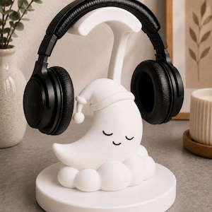 May include: A white headphone stand featuring a sleeping moon design with closed eyes and a nightcap. Black headphones rest on the stand. The stand is on a round base. The Sony headphones are black.