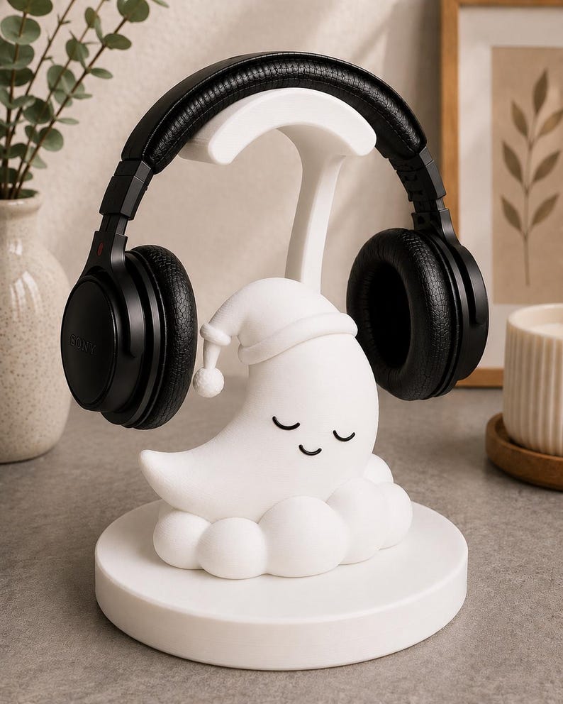 May include: A white headphone stand featuring a sleeping moon design with closed eyes and a nightcap. Black headphones rest on the stand. The stand is on a round base. The Sony headphones are black.