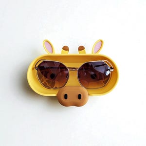 K&ouml;nnte beinhalten: Ein gelber Sonnenbrillenhalter in Form eines Giraffengesichts mit braunen Sonnenbrillen. Der Halter hat eine braune Nase und rosa gef&uuml;tterte Ohren. Die Sonnenbrille hat einen goldenen Rahmen und braune Gl&auml;ser.
