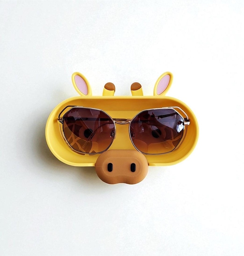 K&ouml;nnte beinhalten: Ein gelber Sonnenbrillenhalter in Form eines Giraffengesichts mit braunen Sonnenbrillen. Der Halter hat eine braune Nase und rosa gef&uuml;tterte Ohren. Die Sonnenbrille hat einen goldenen Rahmen und braune Gl&auml;ser.