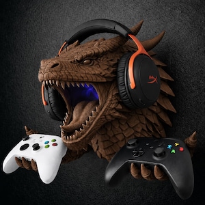 May include: A brown dragon head wall mount featuring textured scales and an open mouth with a blue light effect. The dragon wears black and orange over-ear headphones and has two clawed hands holding a white game controller and a black game controller.