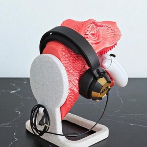 May include: A red dinosaur head-shaped headphone stand with a black headset and a white game controller. The stand is made of a speckled gray material and has a base for stability. The headset has gold accents and the text "NXS".