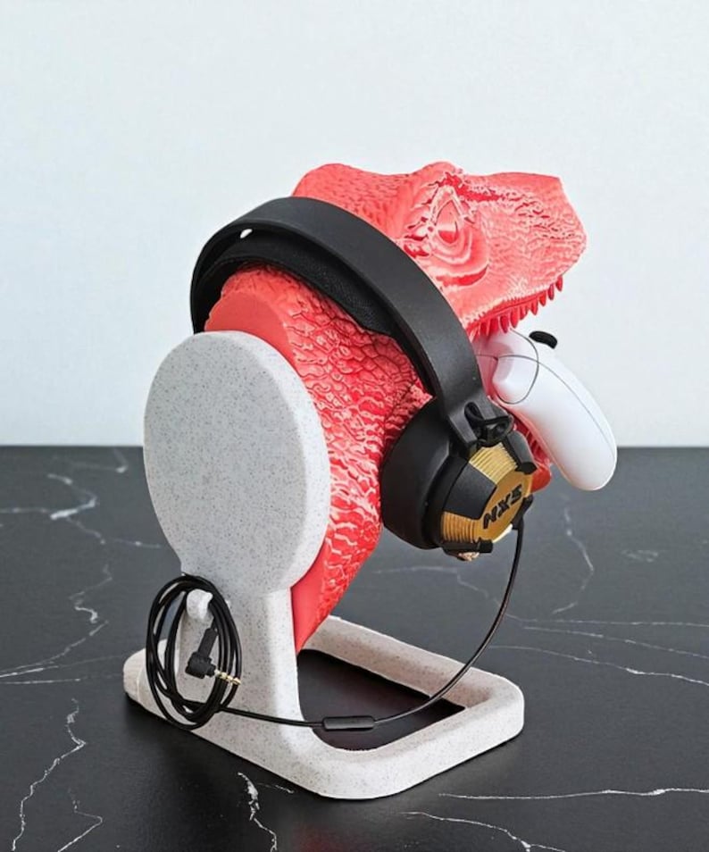 May include: A red dinosaur head-shaped headphone stand with a black headset and a white game controller. The stand is made of a speckled gray material and has a base for stability. The headset has gold accents and the text "NXS".