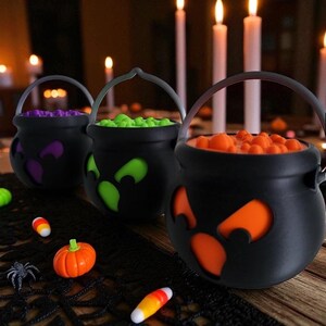 May include: Three black cauldron-shaped Halloween candy holders with cut-out eye designs, each filled with colorful candies. A small plastic pumpkin, spider, and candy corn are scattered nearby. The background features lit candles.