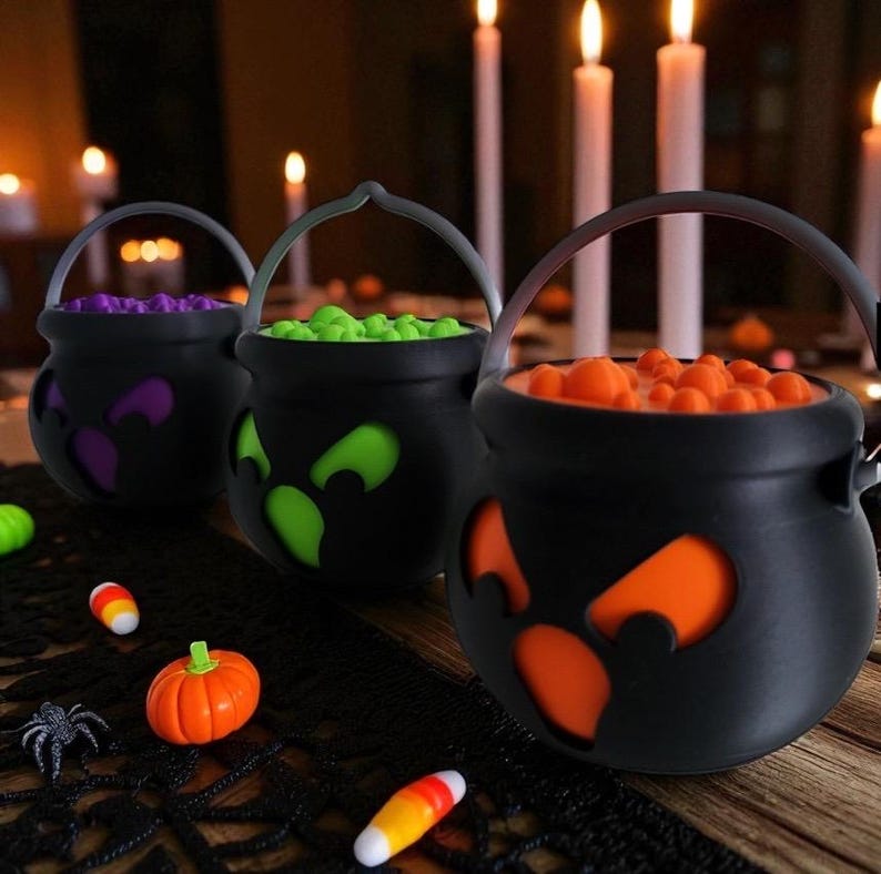 May include: Three black cauldron-shaped Halloween candy holders with cut-out eye designs, each filled with colorful candies. A small plastic pumpkin, spider, and candy corn are scattered nearby. The background features lit candles.