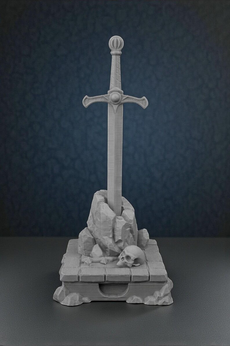 May include: A gray sculpture of a sword in a stone, with a skull at the base. The sword has a detailed hilt and is set in a rocky formation atop a rectangular base. The overall design evokes a fantasy theme.