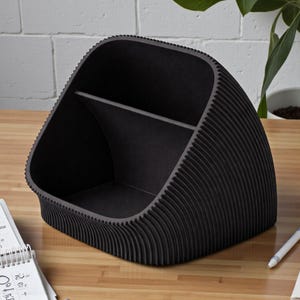 May include: A black desk tidy with a distinctive, wave-like design. The organiser has two compartments and is made of a ridged material. It sits on a wooden surface, with a white brick wall in the background.