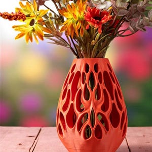 以下が含まれることがあります： 鮮やかなオレンジ色の花瓶。特徴的な透かし彫りのデザインで、人工的な花がカラフルにアレンジされています。花瓶には多数の楕円形の開口部があります。花には、黄色、オレンジ色、赤色の花と、緑と茶色の葉が含まれています。