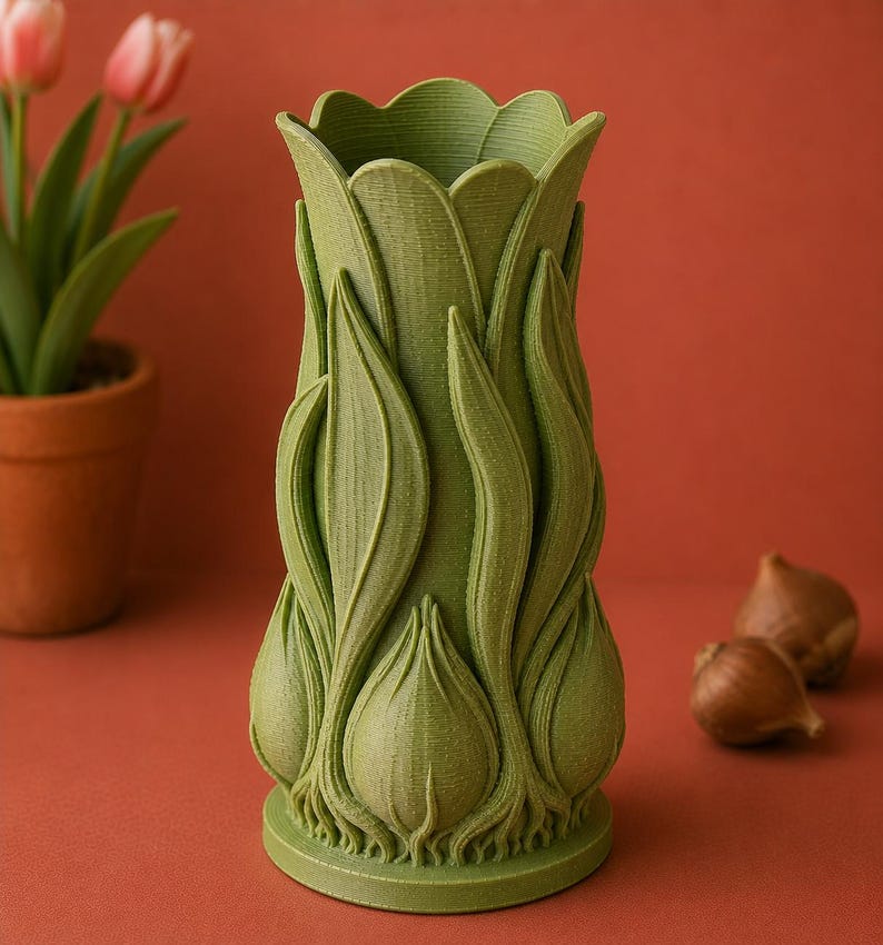 May include: A green, tulip-shaped vase with a textured surface. The vase has a detailed design of tulip leaves and bulbs. The vase is on a red-orange surface, with a potted plant and tulip bulbs in the background.