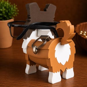 May include: A wooden dog-shaped eyeglasses holder in brown, white, and tan. The dog has a tail and is wearing black eyeglasses. The holder has a metal ring attached to the dog's body. The dog is sitting on a wooden surface.