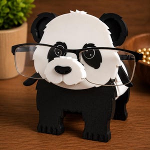 May include: A black and white panda-shaped eyeglasses holder with black-framed glasses. The holder has a white face with black eyes and ears, and a black nose. The panda is standing on four black legs.