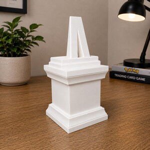 May include: A white, geometric sculpture with a tiered base and a central, inverted V-shaped element. The sculpture is placed on a wooden surface, with a potted plant and a desk lamp in the background. The sculpture is approximately 8 inches tall.