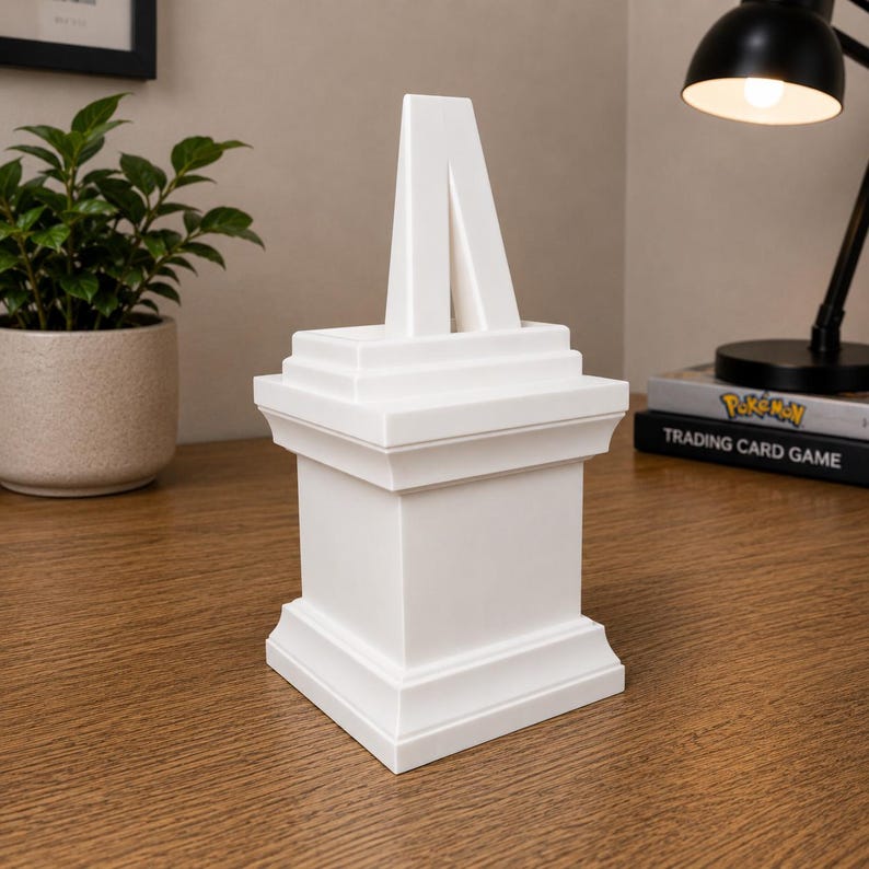 May include: A white, geometric sculpture with a tiered base and a central, inverted V-shaped element. The sculpture is placed on a wooden surface, with a potted plant and a desk lamp in the background. The sculpture is approximately 8 inches tall.