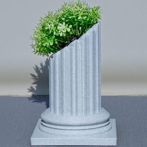 May include: A grey, fluted column planter with a square base, holding a cluster of green plants. The top of the planter is cut at an angle, creating a unique design. The planter is made of a stone-like material.