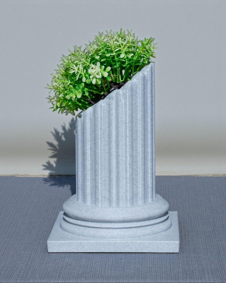 May include: A grey, fluted column planter with a square base, holding a cluster of green plants. The top of the planter is cut at an angle, creating a unique design. The planter is made of a stone-like material.