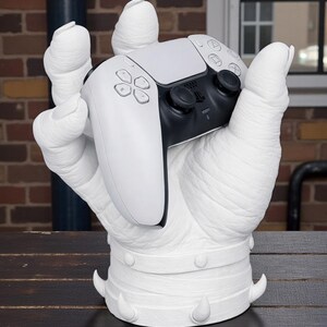 May include: A white sculpture of a hand holding a black and white video game controller. The hand has a textured surface and is mounted on a base with decorative spikes. The controller features buttons and directional controls.