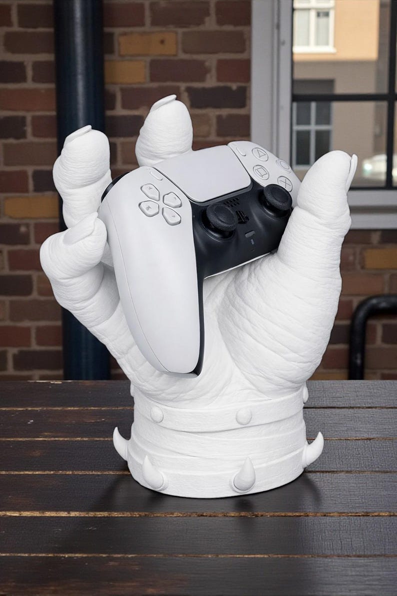 May include: A white sculpture of a hand holding a black and white video game controller. The hand has a textured surface and is mounted on a base with decorative spikes. The controller features buttons and directional controls.