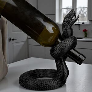May include: A black snake-shaped wine bottle holder. The holder is made of a textured material and is holding a wine bottle. The snake's head is raised, and its mouth is open. The bottle is green.