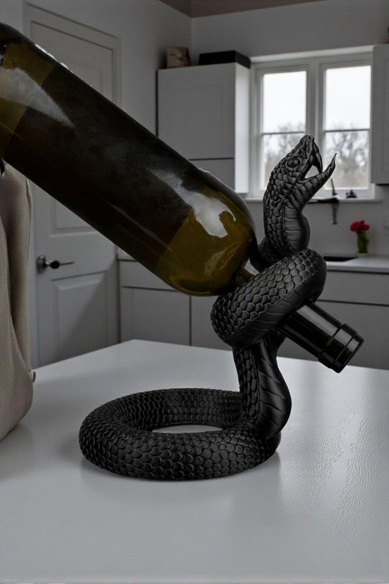 May include: A black snake-shaped wine bottle holder. The holder is made of a textured material and is holding a wine bottle. The snake's head is raised, and its mouth is open. The bottle is green.