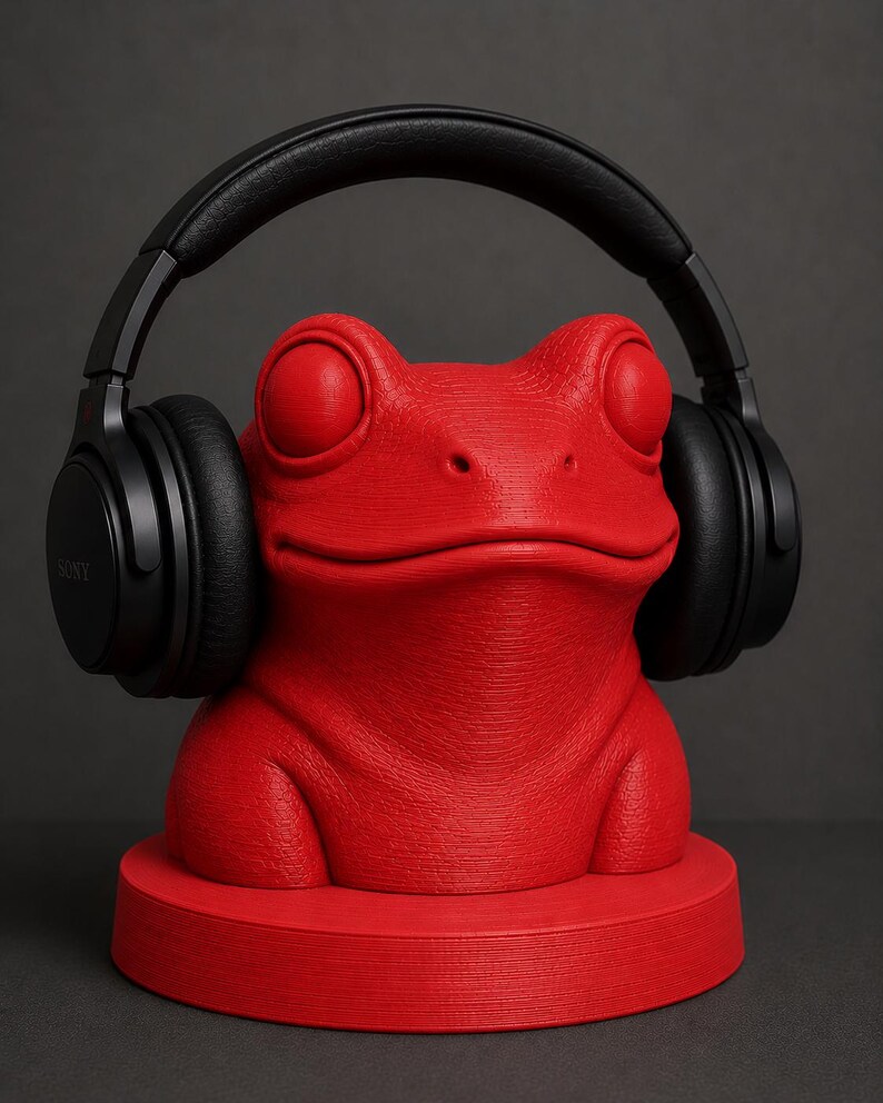 May include: A bright red frog-shaped headphone stand with black headphones. The stand has a textured surface and a round base. The headphones are black and have the brand name "SONY" printed on the side.