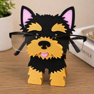 May include: A black, gold, and pink dog-shaped eyeglasses holder. The holder has a textured gold face and paws, black fur, and pink inner ears. Black eyeglasses are resting on the holder. The holder is on a wooden surface.
