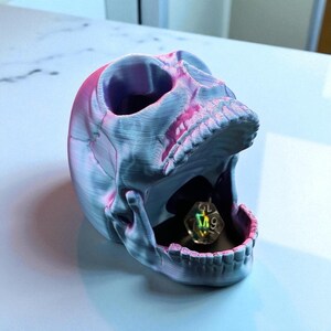 May include: A 3D-printed skull-shaped container in shades of blue and pink, with a multi-sided die inside its open mouth. The skull has a textured surface and detailed teeth, perfect for tabletop gaming enthusiasts.