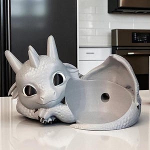 Puede incluir: Una maceta con forma de drag&oacute;n gris claro con grandes ojos negros y una maceta gris a juego. El drag&oacute;n est&aacute; en una pose de descanso, con la cabeza y las patas apoyadas en una superficie. La maceta est&aacute; abierta y lista para plantar.