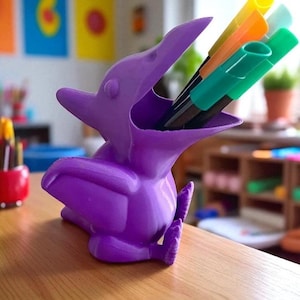 May include: A purple pterodactyl-shaped desk organizer holding colorful markers. The organizer is made of plastic and sits on a wooden surface. The markers are yellow, orange, and green. The background includes colorful artwork.