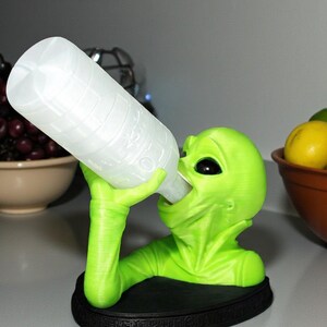 May include: A neon green alien figurine holding a white bottle. The alien is drinking from the bottle, which is angled towards its mouth. The figurine sits on a black base. The alien has large black eyes and a smooth texture.
