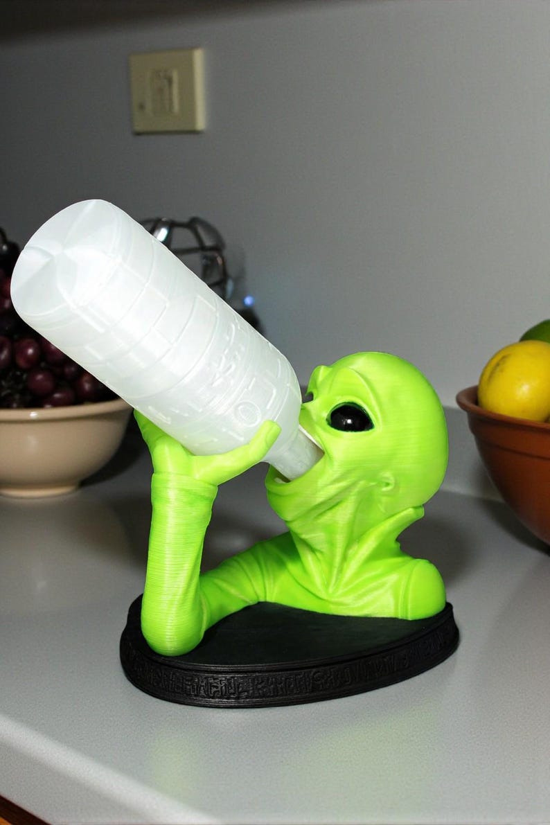 May include: A neon green alien figurine holding a white bottle. The alien is drinking from the bottle, which is angled towards its mouth. The figurine sits on a black base. The alien has large black eyes and a smooth texture.