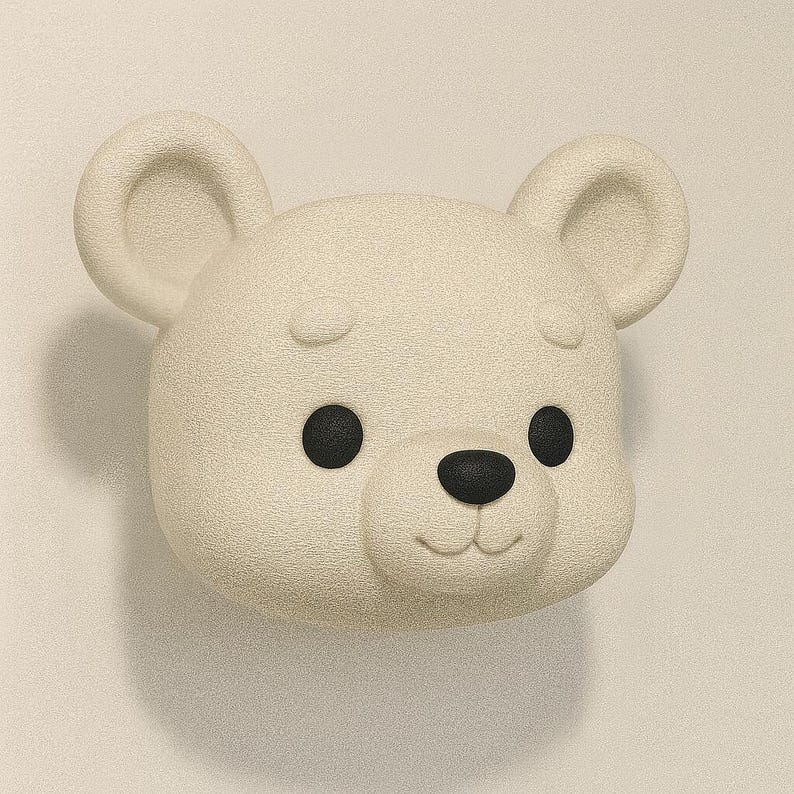May include: A cream-colored bear head with black eyes and nose. The bear has rounded ears and a textured surface. The design is simple and cartoonish, with a smiling expression. The bear is a decorative item.