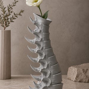 May include: A gray, sculptural vase with a unique, spine-like design, holding a white flower with a green stem and leaf. The vase has a textured surface and is placed on a neutral-toned surface, creating a modern, artistic display.