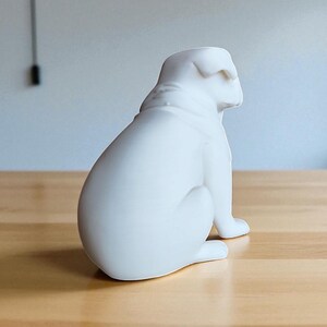 May include: A white ceramic bulldog figurine, sitting upright with its head turned to the right. The sculpture has a smooth, matte finish and is placed on a light-colored wooden surface. The background is a plain wall.