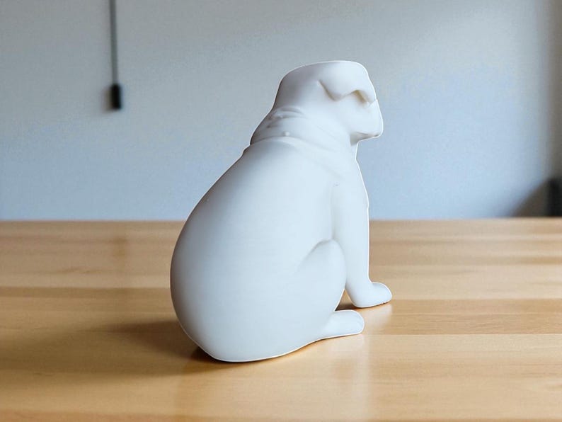 May include: A white ceramic bulldog figurine, sitting upright with its head turned to the right. The sculpture has a smooth, matte finish and is placed on a light-colored wooden surface. The background is a plain wall.