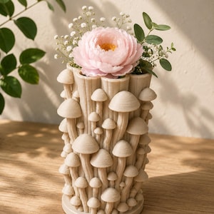 K&ouml;nnte beinhalten: Eine beige Vase mit Pilz-Design h&auml;lt eine rosa Pfingstrose und kleine wei&szlig;e Bl&uuml;ten. Die zylindrische Vase ist mit verschiedenen Pilzformen verziert. Gr&uuml;ne Bl&auml;tter erg&auml;nzen das Blumenarrangement. Die Vase steht auf einer Holzoberfl&auml;che.