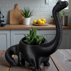 May include: A black dinosaur-shaped plant pot containing a green succulent. The pot is made of a textured material and is on a wooden table. The dinosaur has a long neck and a detailed head.