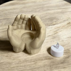 Puede incluir: Un portavelas beige en forma de manos con una hendidura circular en el centro. A la derecha del soporte hay una peque&ntilde;a vela de t&eacute; LED blanca con una parte superior transparente en forma de llama. El soporte est&aacute; sobre una superficie de madera marr&oacute;n claro.