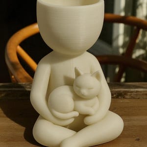 May include: A cream-coloured figurine of a person holding a sleeping cat. The figure has a large head with a hollowed-out top, suggesting a planter or vase. The cat is curled up in the person's lap, creating a serene and comforting scene.