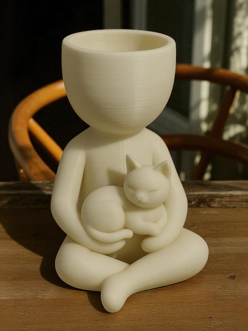 May include: A cream-coloured figurine of a person holding a sleeping cat. The figure has a large head with a hollowed-out top, suggesting a planter or vase. The cat is curled up in the person's lap, creating a serene and comforting scene.
