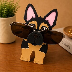 Pode incluir: Um suporte de &oacute;culos de novidade em forma de c&atilde;o pastor alem&atilde;o. O suporte em forma de c&atilde;o preto, castanho e rosa tem &oacute;culos de sol apoiados no nariz. Os &oacute;culos de sol t&ecirc;m arma&ccedil;&atilde;o prateada e lentes castanhas. Uma pequena planta em vaso e um livro est&atilde;o no fundo.