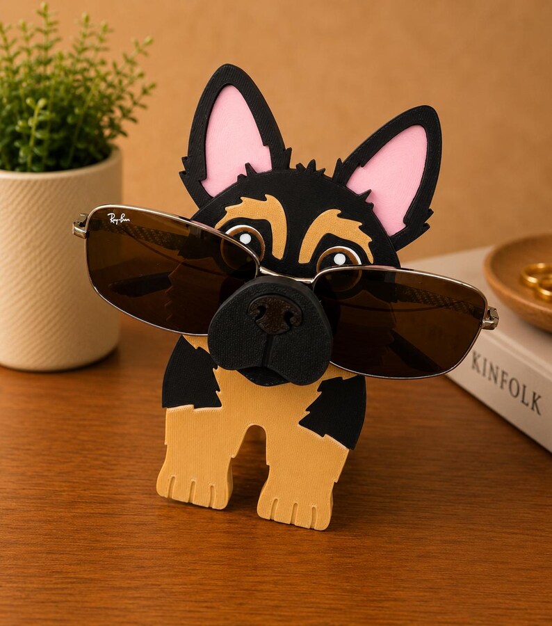 Pode incluir: Um suporte de &oacute;culos de novidade em forma de c&atilde;o pastor alem&atilde;o. O suporte em forma de c&atilde;o preto, castanho e rosa tem &oacute;culos de sol apoiados no nariz. Os &oacute;culos de sol t&ecirc;m arma&ccedil;&atilde;o prateada e lentes castanhas. Uma pequena planta em vaso e um livro est&atilde;o no fundo.