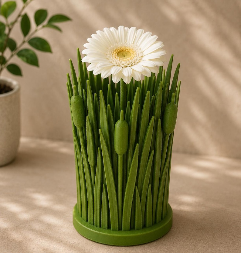 May include: A green, grass-like vase holds a white gerbera daisy with a yellow center. The vase has a cylindrical shape with a round base and is designed to resemble blades of grass and cattails. The background is a neutral color.