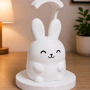 May include: A white bunny-shaped desk lamp with a curved top and a smiling face. The lamp sits on a round base and is made of a smooth, matte material. The lamp is designed to provide soft lighting.