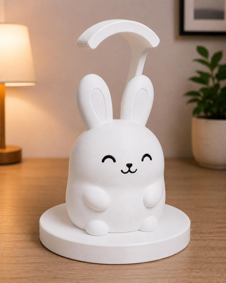 May include: A white bunny-shaped desk lamp with a curved top and a smiling face. The lamp sits on a round base and is made of a smooth, matte material. The lamp is designed to provide soft lighting.