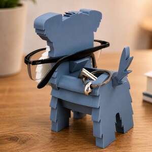 May include: A blue dog-shaped eyeglasses and key holder. The holder is made of layered material and has a slot for glasses and a space for keys. Black-framed glasses and a set of keys are placed in the holder.