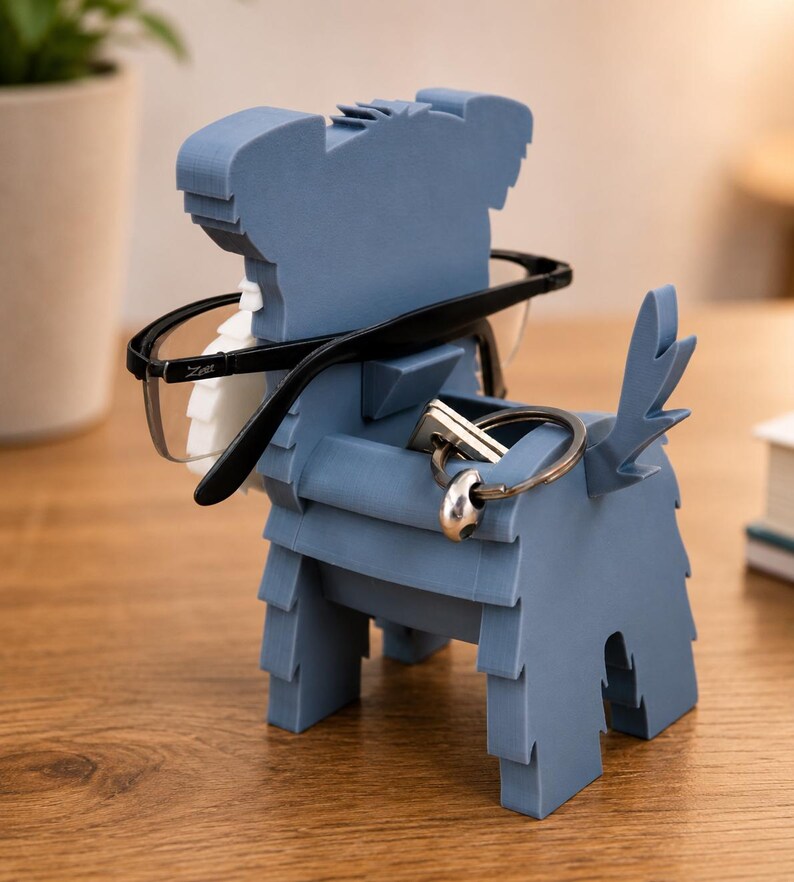 May include: A blue dog-shaped eyeglasses and key holder. The holder is made of layered material and has a slot for glasses and a space for keys. Black-framed glasses and a set of keys are placed in the holder.